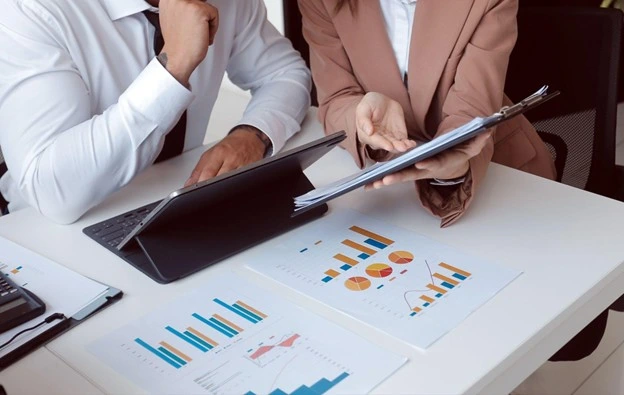 Two business professionals review financial charts and graphs at desk with laptop, collaborating on strategy.