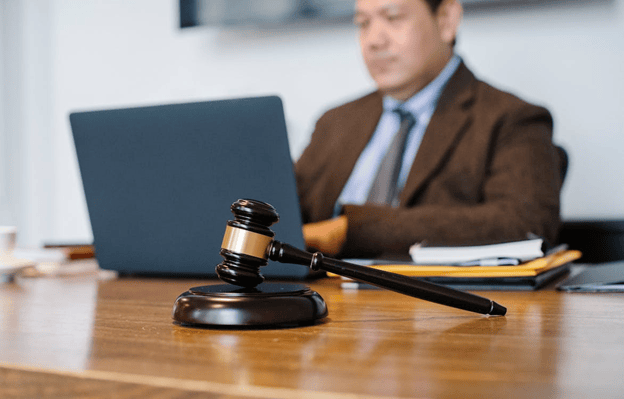 A wooden judge's gavel on a desk with a man in a suit working on a laptop in the background.