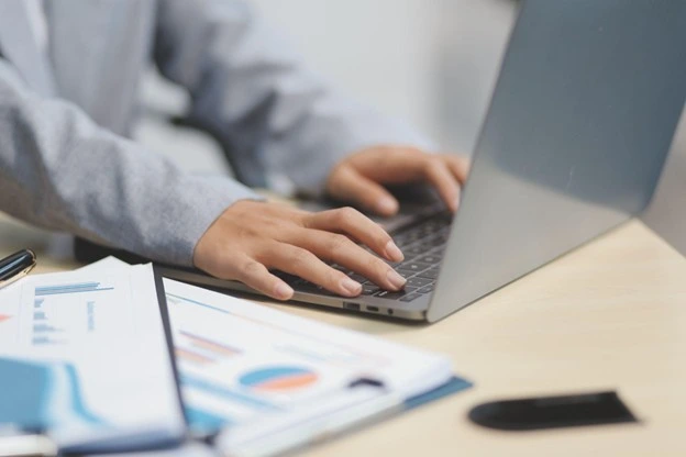 Person in grey suit types on laptop with business charts nearby, actively engaging with data.