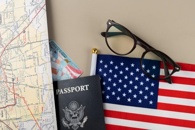 Overhead view of a passport, U.S. flag, glasses, and a map on a tan surface.