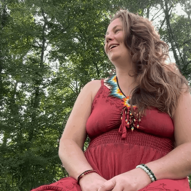 Beautiful woman practicing mindfulness outdoors in a forest setting with lush green trees, wearing a red bohemian dress and colorful beaded necklace, celebrating spiritual wellness and self-care at Soulstice Living.