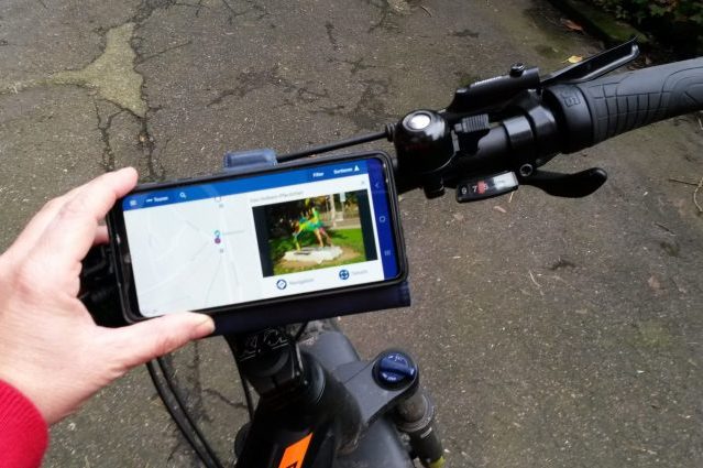 Ein Smartphone am Fahrradlenker zeigt eine App für eine Stadtführung Freiburg mit digitaler Karte und Bild eines Denkmals zur Orientierung an.