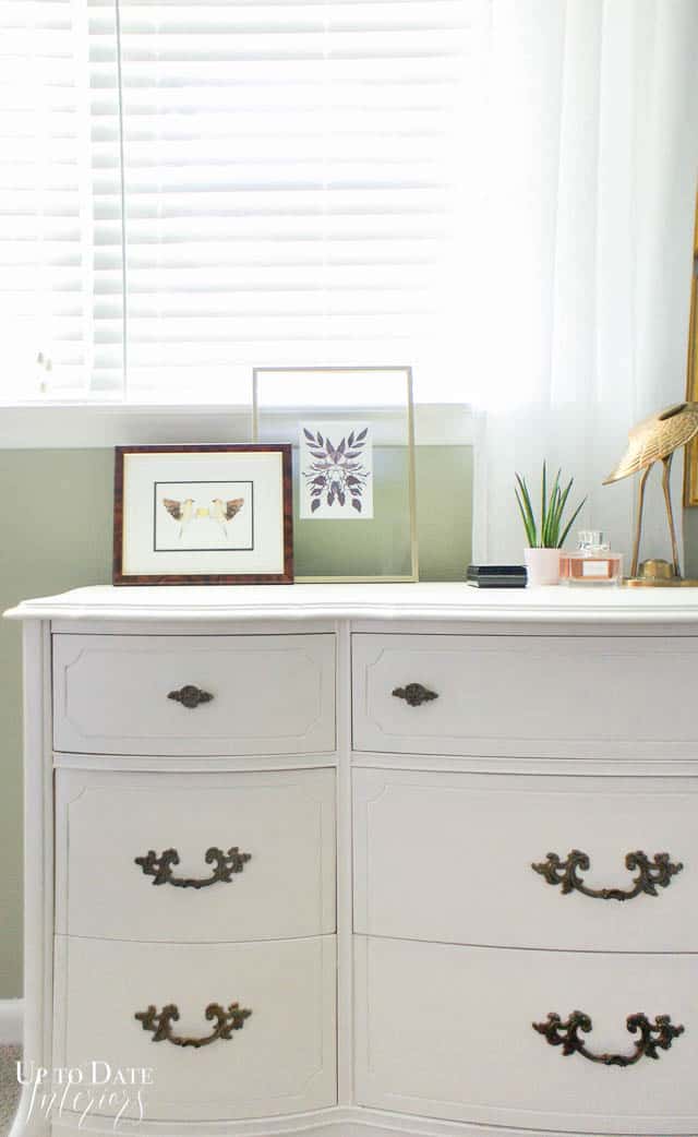 part of white dresser with art and a little plant and brass bird on top. Khaki green wall, white window treatment