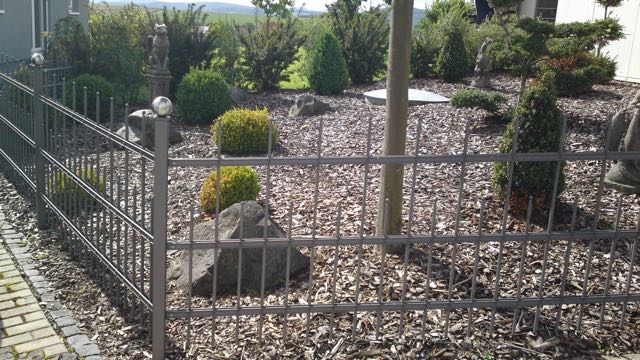 Robuster, anthrazitfarbener Doppelstabmattenzaun mit Kugel-Pfostenkappen von Zaunbau von Stahl- und Metallbau Drost, der einen modern gestalteten Garten mit Rindenmulch, Sträuchern und Dekosteinen eingrenzt.