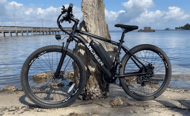 Side profile of an Ancheer Gladiator electric mountain bike near a seawall, highlighting front suspension fork and drivetrain.