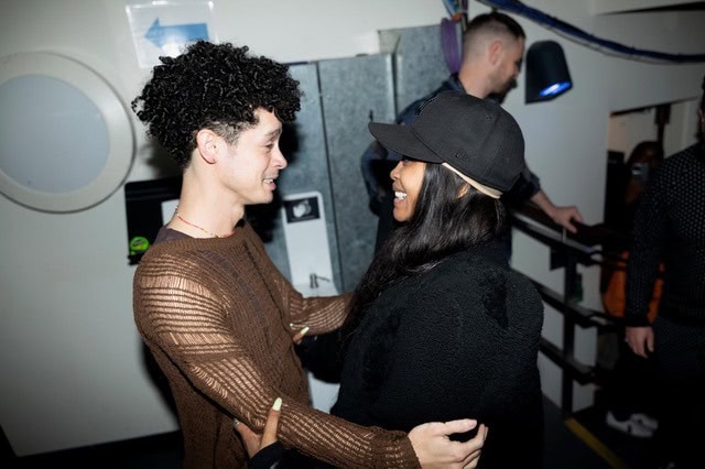 Two people smile at each other and embrace in a warmly lit indoor setting, with another person standing in the background and a circular window on the wall nearby.