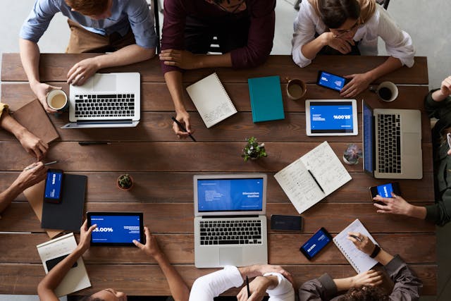 A group of professionals with laptops, tablets, phones, and notebooks meeting at a wooden brown table.