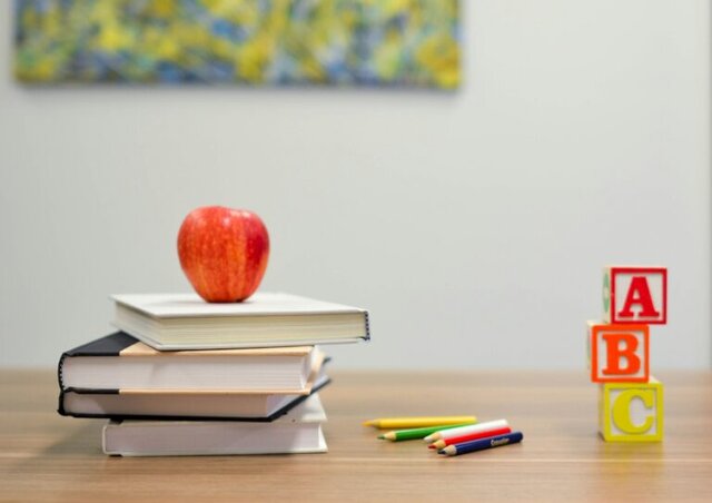 An Apple on top of four books on top of a desk. Colored pencils and a mug accompany them.