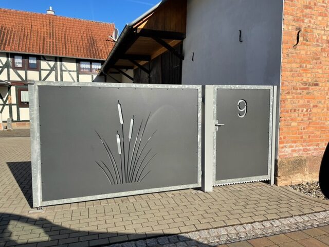 Zweigeteiltes dunkelgraues Metalltor von Stahl- und Metallbau Drost, links mit einem lasergeschnittenen Schilf-Motiv, rechts eine Fußgängertür mit Hausnummer 9. Das Tor hat einen verzinkten Rahmen.