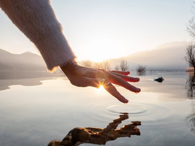 Detail of hand touching and caressing water surface of beautiful lake at sunset, mountain view. Purity freshness clean concept, one person touching lake with hand