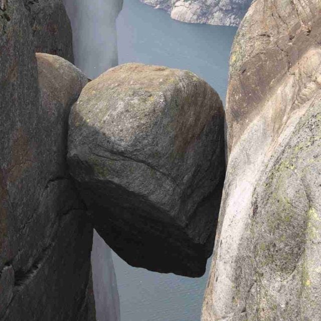 A large boulder is wedged between two cliffs above a body of water, with rocky surfaces visible on all sides.