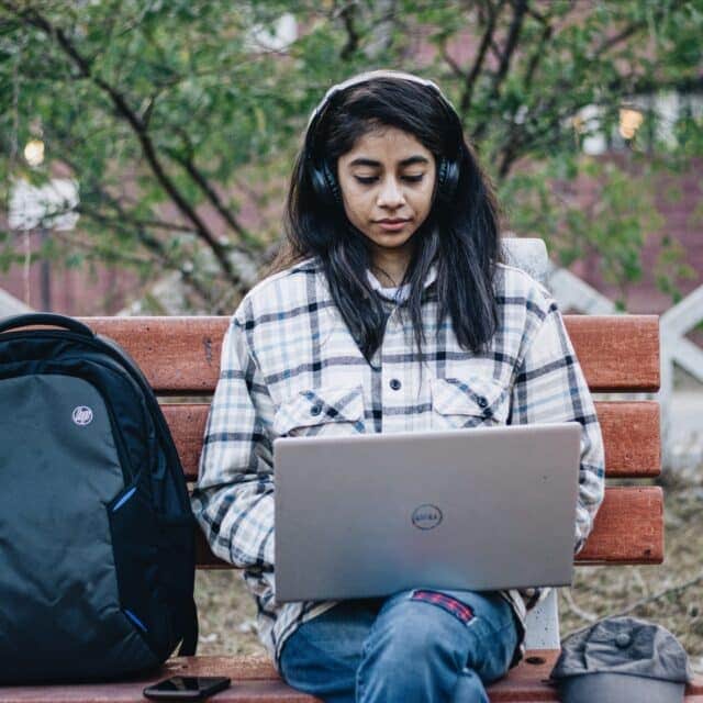 A girl sits on a bench on laptop and headphones having a telehealth appointment with a Clinical Psychologist