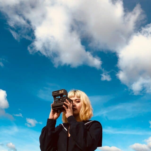 A girl of asian descent with blonde hair takes a polaroid with a blue sky background behind her