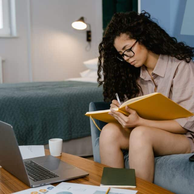 A girl sees a Clinical Psychologist remotely on her laptop and takes notes in a journal, while sitting on a couch