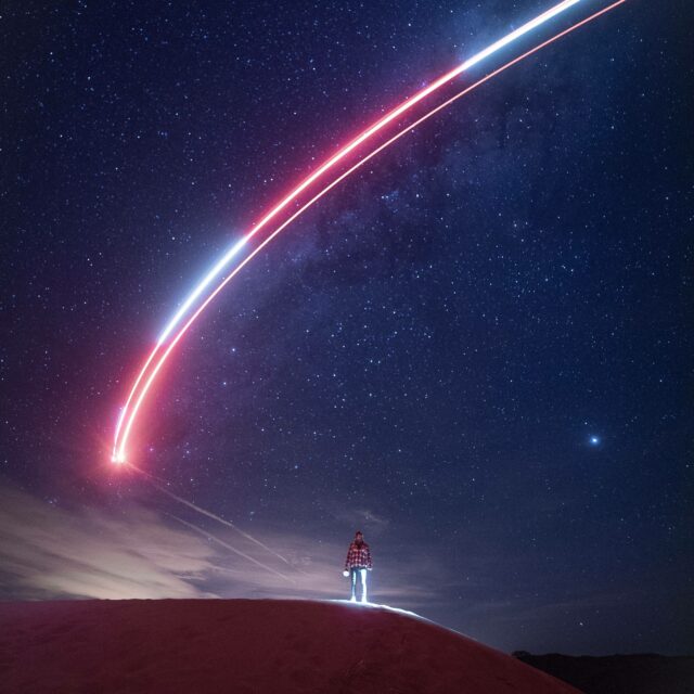 A long exposure coloured streak through the night sky with a figure standing beneath it