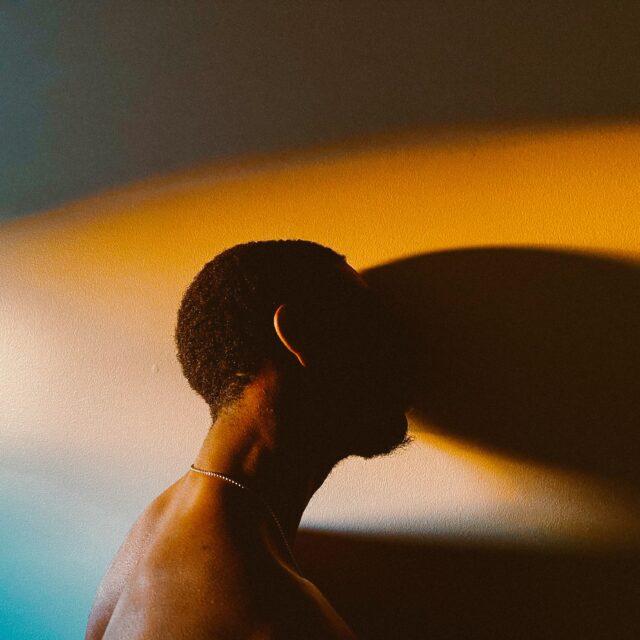 A man rests his head against the wall, his face is obscured by shadows as coloured lights of orange and blue light the wall.