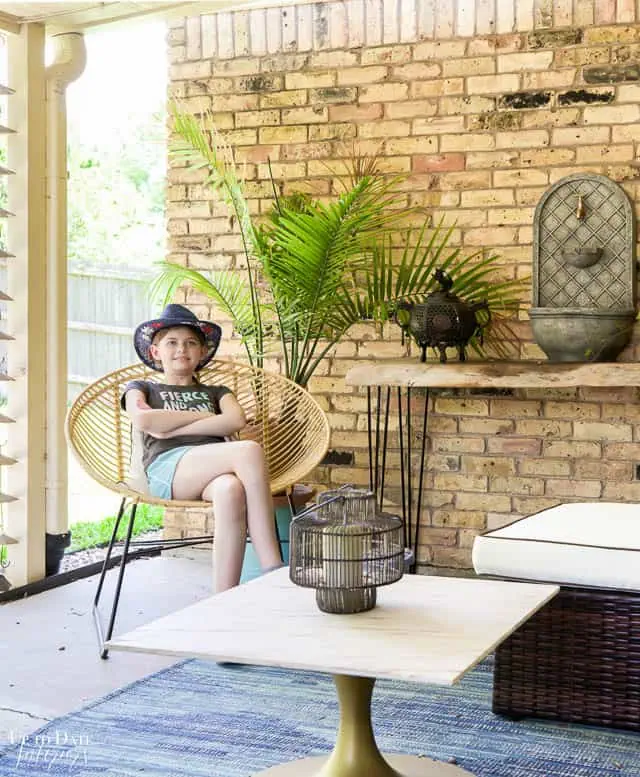 View of outdoor living room with girl sitting in a hair, palm tree, table with fountain and pale yellow bricks