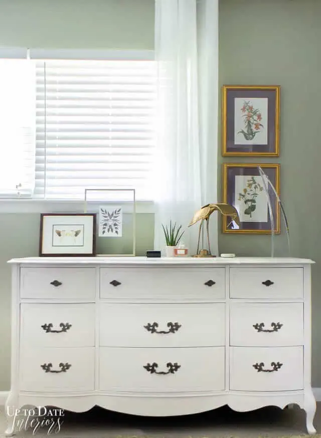 How to paint a dresser white! White French provincial style dresser against a window and green khaki walls with botanical art prints and decor styled on top of dresser including a brass crane and lucite sculpture.
