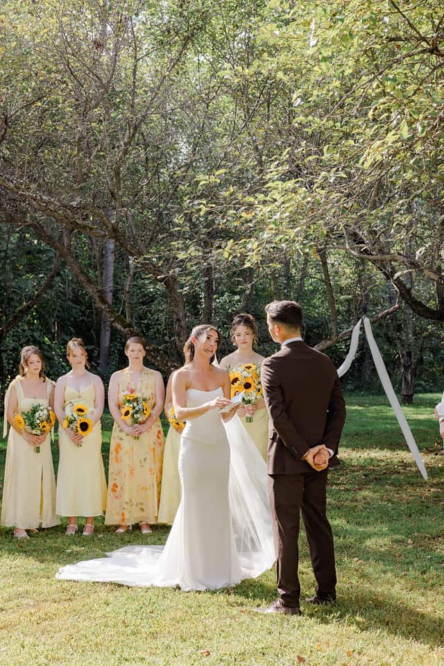 Wide shot of the wedding ceremony with bridesmaids looking on as Julia and Will exchange rings under the trees