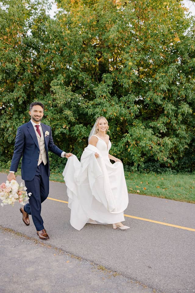 Bride and groom laughing as they walk hand in hand down a tree lined path