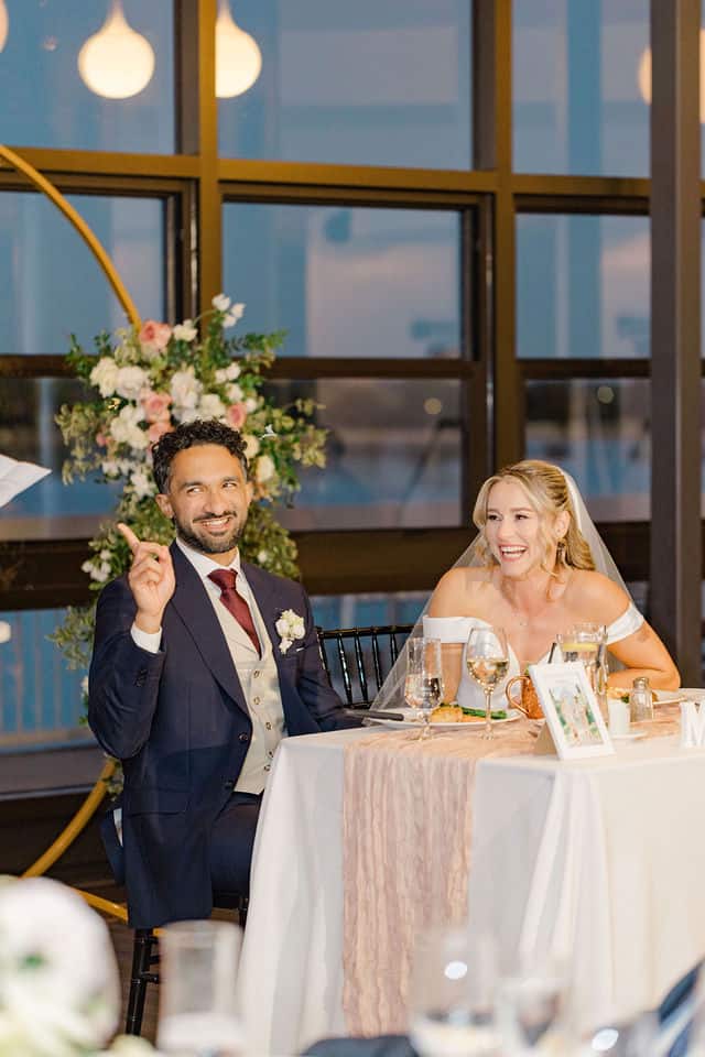 Groom mid toast at the sweetheart table as the bride laughs beside him