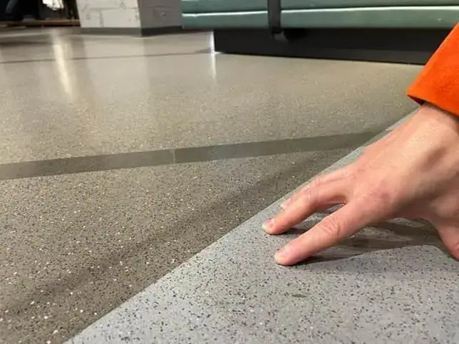 Dettaglio ravvicinato di un pavimento in resina Ultratop Polished con finitura granulata, realizzato in due varianti cromatiche — mid gray e dark gray — accostate con una giunzione netta. Una mano sfiora la superficie per mostrarne la texture e la qualità della posa, in un ambiente retail.