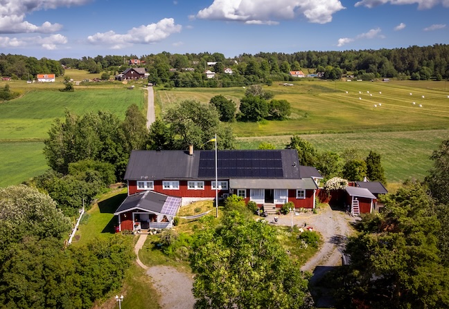 Flygfoto över ett rött hus med svarta solceller på taket, omgivet av grönska, grusväg och öppna fält under en blå himmel.