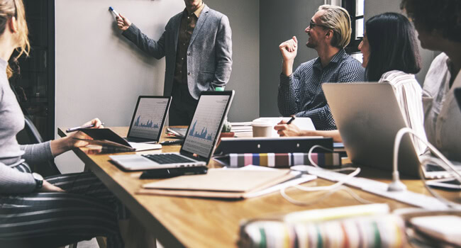 Business team in a meeting, with people around a table, laptops open, and one person presenting on a whiteboard.