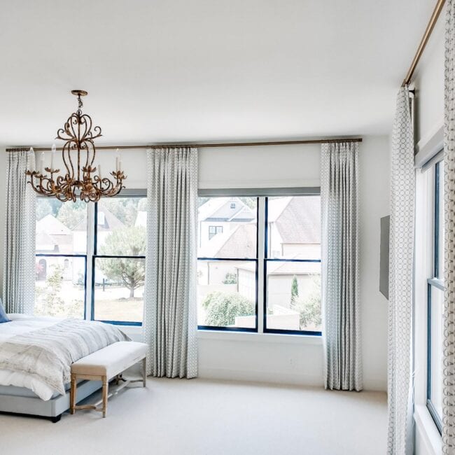 Sunny bedroom featuring expansive windows, stylish patterned curtains, and a luxurious chandelier.