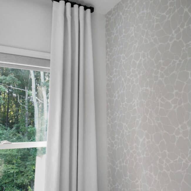 White curtain hanging indoors near large window with greenery outside.