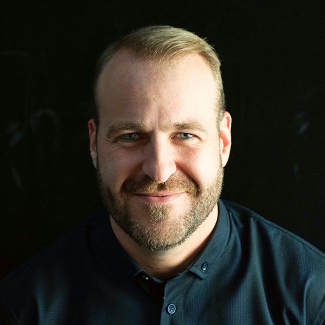 Modern architect portrait of a man with a beard and blue eyes, against a dark background.
