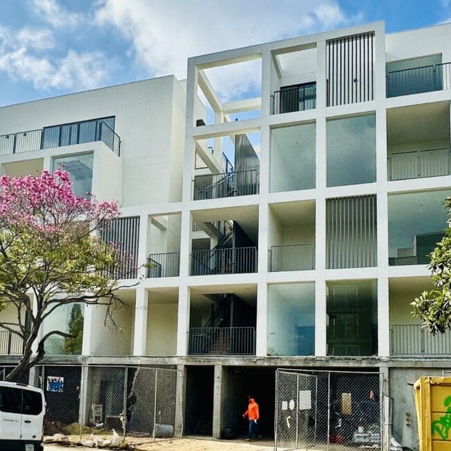 Modern white multi-story apartment building with open-air corridors and balconies, featuring vertical slat accents and a geometric facade.