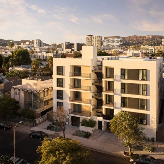 Modern white apartment building with balconies and large windows, set in an urban neighborhood at sunset.