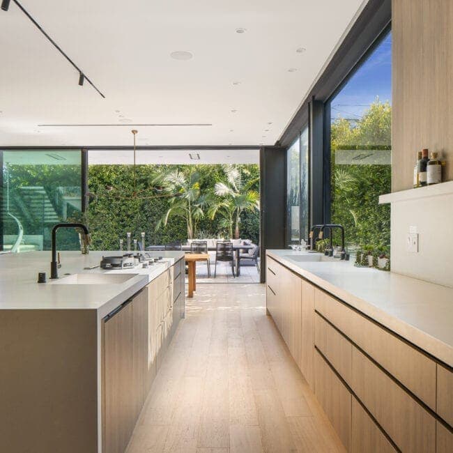 Contemporary kitchen with minimalist design, white surfaces, natural wood cabinetry, expansive glass sliding doors showing lush greenery outside.