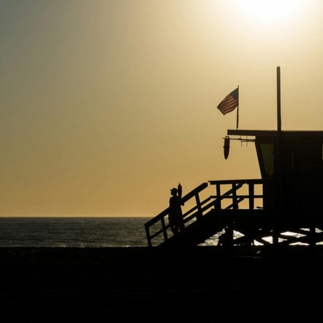 Silhouette of a lifeguard tower with an American flag on a beach during sunset.