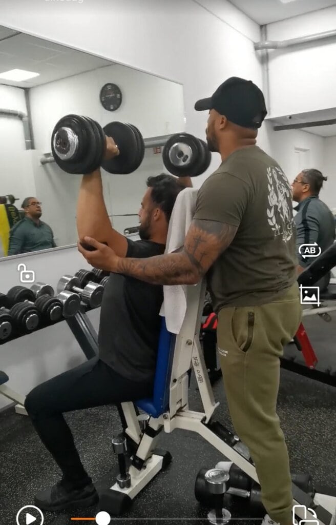 Sporters ondersteunten elkaar tijdens een dumbbell shoulder press in de sportschool, met aandacht voor correcte techniek en veiligheid