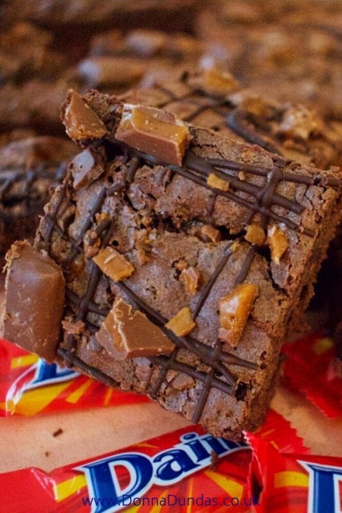 Square of brownie on side, topped with broken daim bars. More brownie blurred in background, Daim Bars in wrappers in foreground