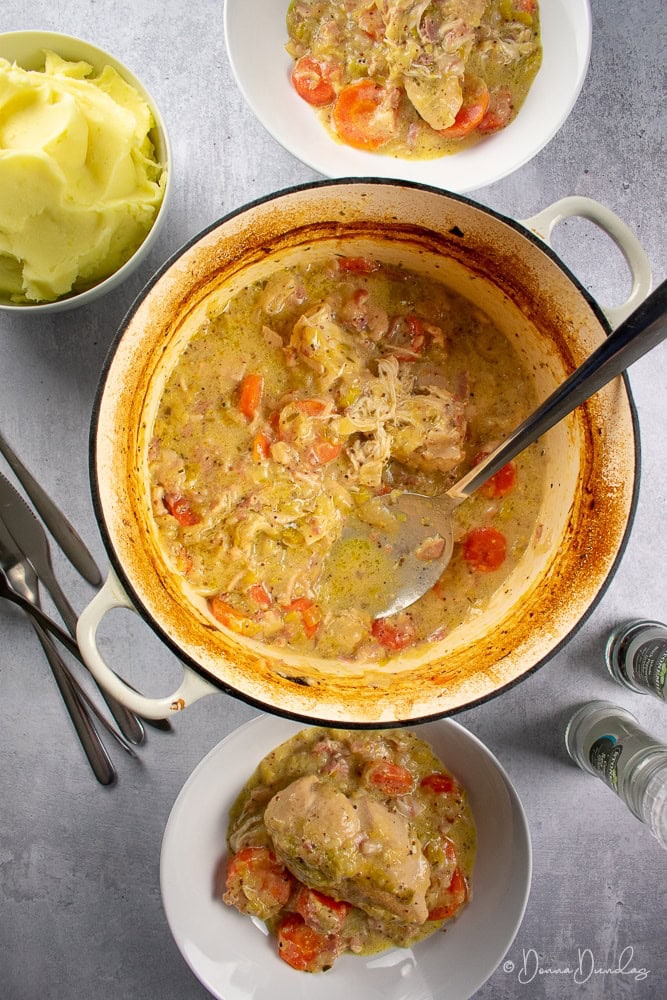 Cast iron dish with chicken casserole in middle, 2 served portions top and bottom, mashed potato and cutlery on table
