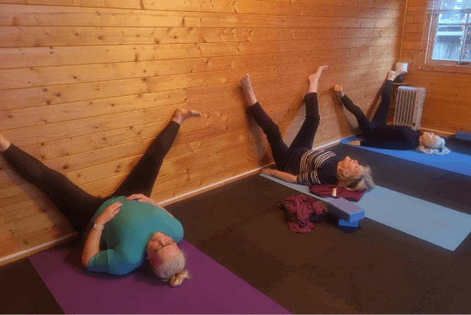 Women in a Croxley Green yoga class lying on mats with legs supported against a wooden wall during a gentle stretch.