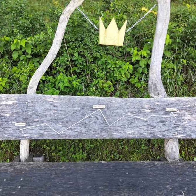 Sich wiederholende Bank auf dem König-Ludwig-Weg. Auf der Rückenlehne ist der Weg aufgezeichnet. Über der Bank ist an zwei Stangen eine goldene Krone angebracht.