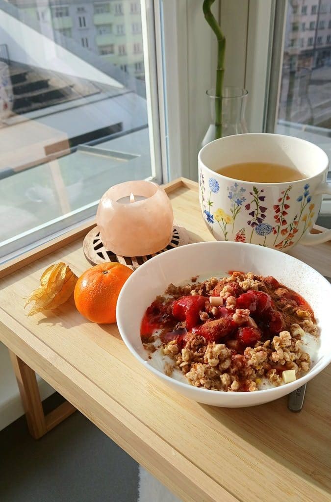 Eine Joghurt-Müsli-Bowl mit Beeren und eine Tasse Tee auf einem Holztablett am Fenster.