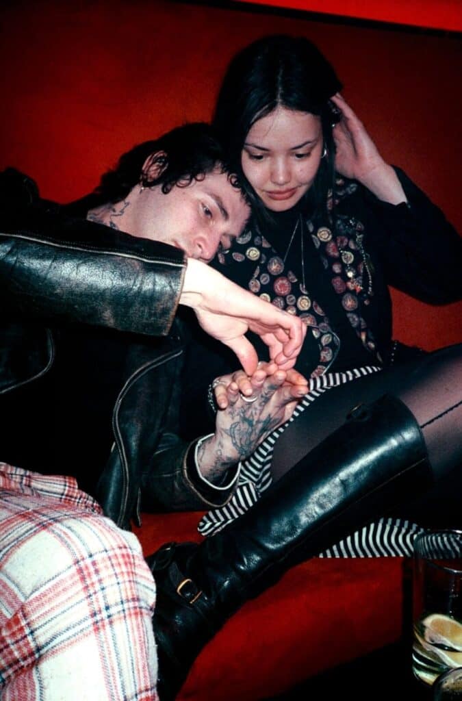 Two young people sitting on a red velvet sofa in a dimly lit venue, the man in a leather jacket resting his head on the woman's shoulder as they hold hands — photograph by Arthur Samier