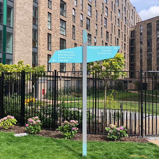 Fingerpost sign with a grey post with two light blue paddle signs attached towards the top and right angles to the post. These feature directional information about key destinations in white lettering. There is blue decoration around the top and bottom of the post and the sign is installed in a grassy area in front of an apartment building