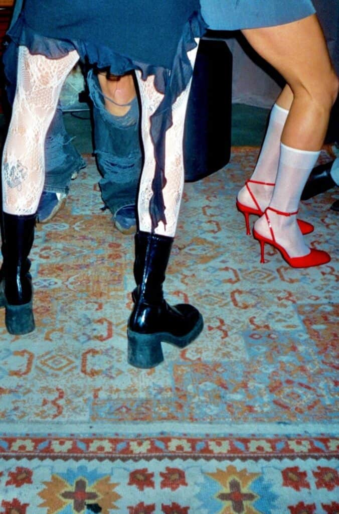 Detail shot of three pairs of legs on a patterned rug: lace tights with black platform boots, ripped jeans, and white socks with red strappy stilettos — photograph by Arthur Samier
