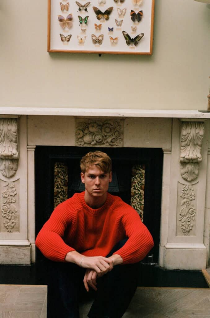 Dan Whitlam, a man with short hair in a bright red sweater, sits on the floor in front of an ornate white fireplace, with a framed display of butterflies hanging above the mantel.