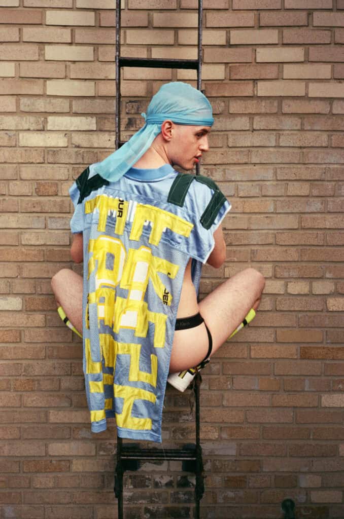 A figure wearing a blue durag and a pale blue and yellow mesh sports jersey climbing a black metal ladder against a brick wall, Calvin Klein underwear visible — photograph by Slava Mogutin