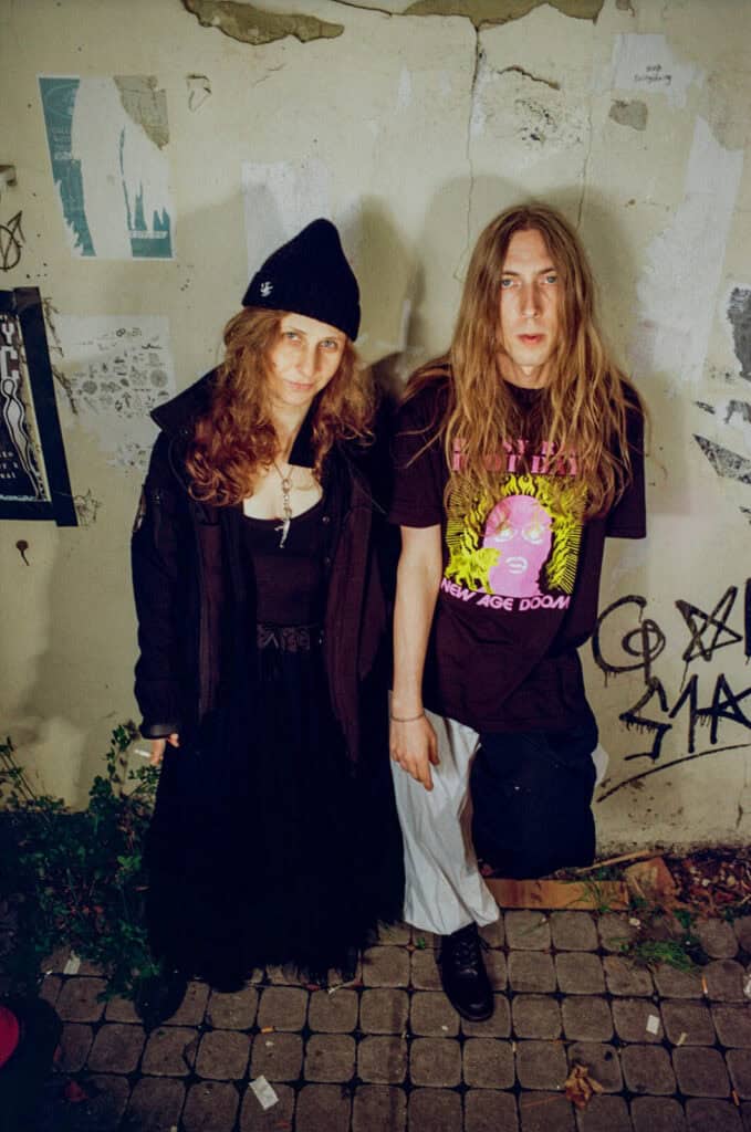 Maria Alyokhina of Pussy Riot and a guy stand together in front of a weathered wall with graffiti and torn posters. One, wears a black outfit and beanie; the other has long hair and a graphic T-shirt. Both look at the camera with neutral expressions.