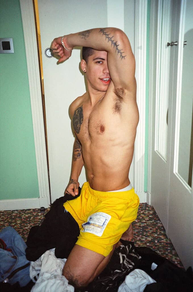 A tattooed shirtless young man in yellow shorts kneeling on a hotel room floor amid scattered clothing, one arm raised above his head, smiling — photograph by Slava Mogutin