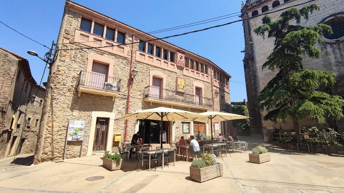 Old stone building with outdoor cafe seating, umbrellas, and pedestrians in a historic European town square.