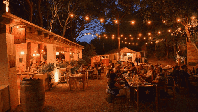 Cozy outdoor dining at Can Camades restaurant during evening with string lights, wooden tables, and lush trees creating a warm, inviting atmosphere for guests.